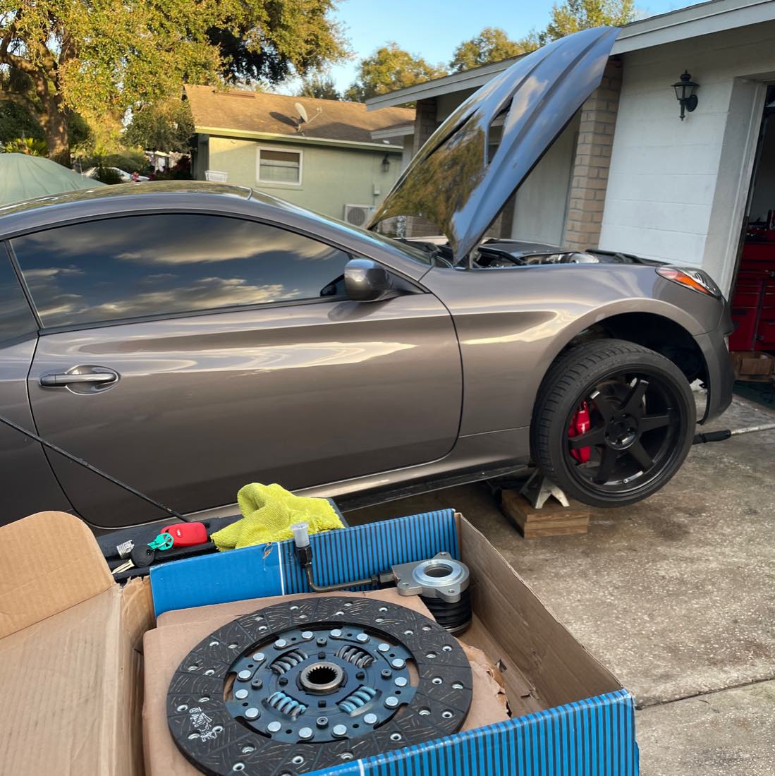 Pat's Mobile Auto Repair replacing a clutch on a coupe in a customer's driveway, new clutch kit staged in the foreground